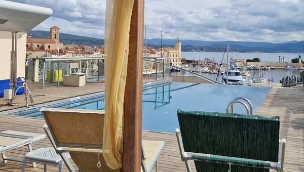 Blick von einem Loungebett auf den Pool und im Hintergrund der Hafen und das Meer.
