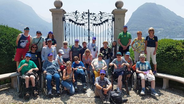 Eine Gruppe von Menschen posiert vor einem grossen Tor und im Hintergrund ist ein See.