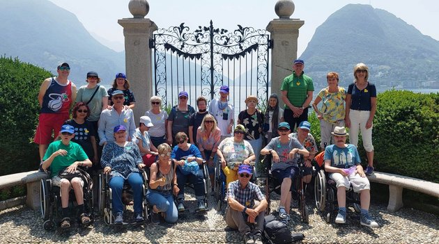 Eine Gruppe von Menschen posiert vor einem grossen Tor und im Hintergrund ist ein See.