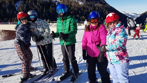 Eine Gruppe von fünf Skifahrer*innen steht bei strahlendem Sonnenschein am Pistenrand und lächelt in die Kamera.
