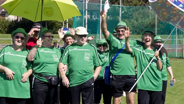 Eine Gruppe posiert auf einem Sportplatz. Alle haben grüne Tshirts an und lachen in die Kamera. 