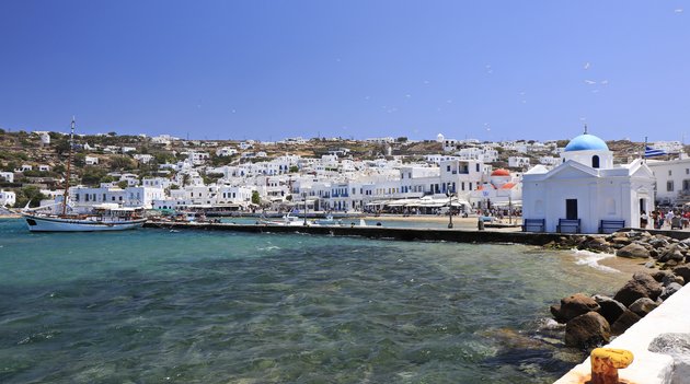 Im Vordergrund das tosende Meer und im Hintergrund die typischen griechischen Häuser auf der Insel Mykonos.