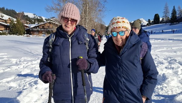 Zwei Frauen dick eingepackt spazieren bei Sonnenschein einem ebenen Winterwanderweg entlang.