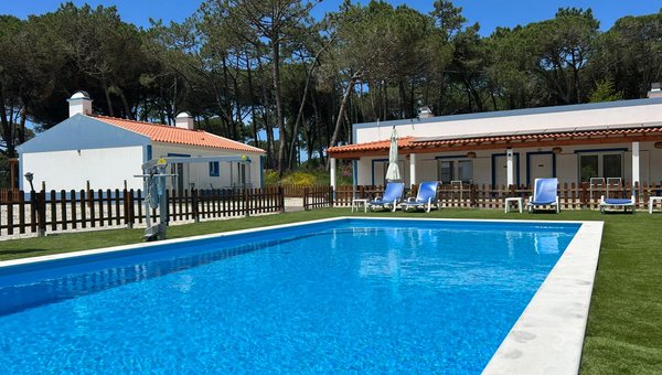 Blick auf den Pool mit Poollift umgeben von ein paar Liegestühlen und im Hintergrund zwei Bungalows und Bäume