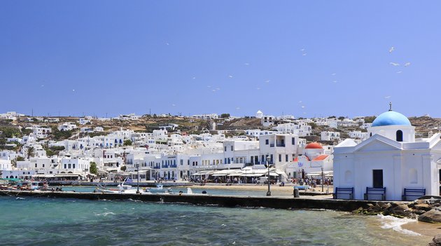 Im Vordergrund das tosende Meer und im Hintergrund die typischen griechischen Häuser auf der Insel Mykonos.