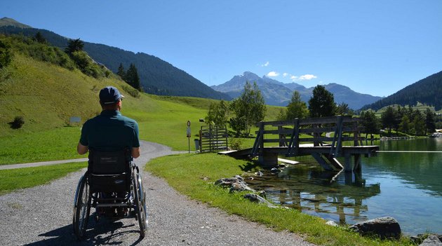Mann im Rollstuhl beim Wandern an einem See und vor einer Bergkulisse