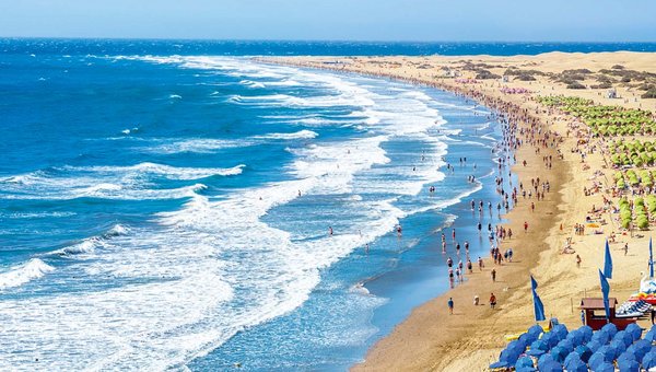 Gigantischer Sandstrand mit aufgewühltem Meer bei schönem Wetter. Im Vordergrund einige Reihen mit blaue Sonnenschirme, im Hintergrund gelbe Sonnenschirme