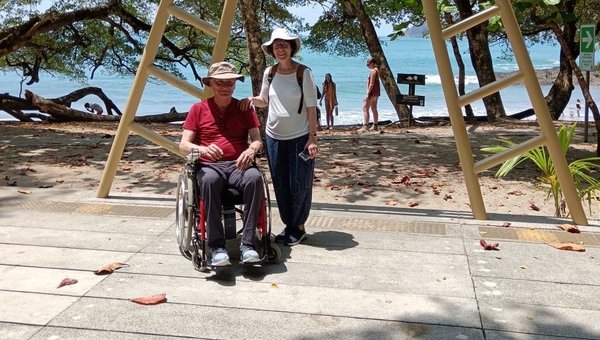 Zwei Touristen, eine Fussgängerin und ein Rollstuhlfahrer posieren vor dem Schild Manuel Antonio Nationalpark. Im Hintergrund ist das Meer mit Sandstrand