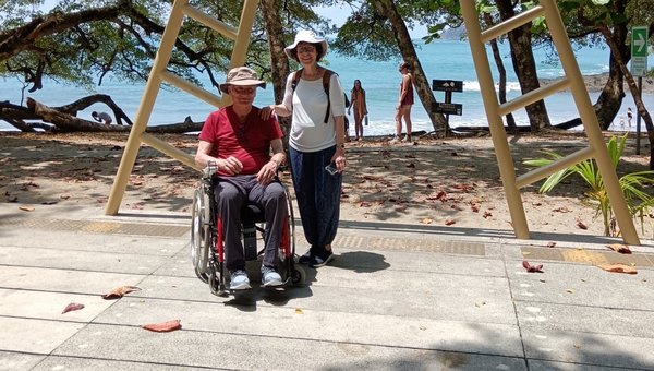 Zwei Touristen, eine Fussgängerin und ein Rollstuhlfahrer posieren vor dem Schild Manuel Antonio Nationalpark. Im Hintergrund ist das Meer mit Sandstrand